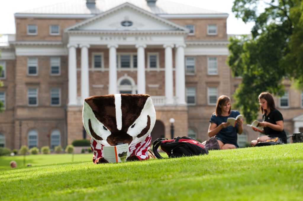 Bucky Badger reading a book on the lawn in front of Bascom Hall