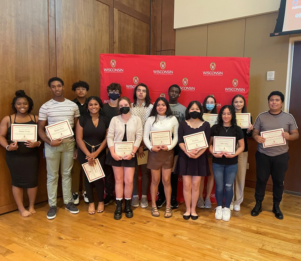 A group of young students stands together holding their earned certificates in front of a UW display.