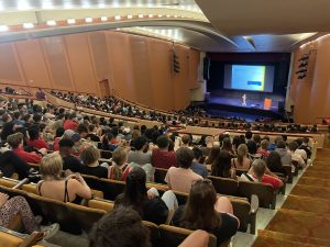 View from the balcony of Shannon Hall down to the stage where a single presenter stands and the seats are full of people.