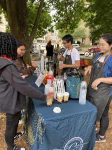 Two people stand behind a table serving different boba tea options to two patrons.