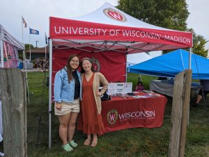 Two HMong women stand in front of a tent and table for UW-Madison.