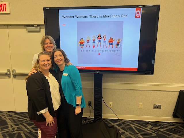 Three women stand in front of a monitor.