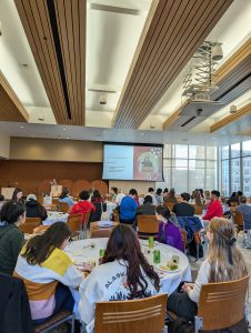 A room filled with students sitting and round tables listening to a presentation
