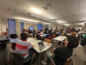 A large group of students sit around tables watching a women give a presentation with a TV screen behind her.