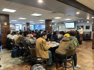 A large group of students sit around tables, eating, and watching a women give a presentation with a TV screen behind her.