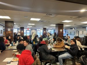 A large group of students sit around tables, eating, and watching a women give a presentation with a TV screen behind her.