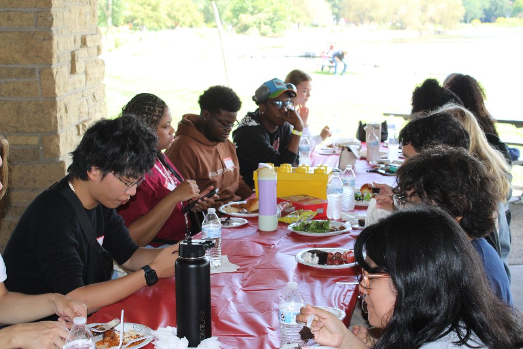 People seated at a long outdoor table with food and drinks, enjoying a communal meal in a picnic setting.