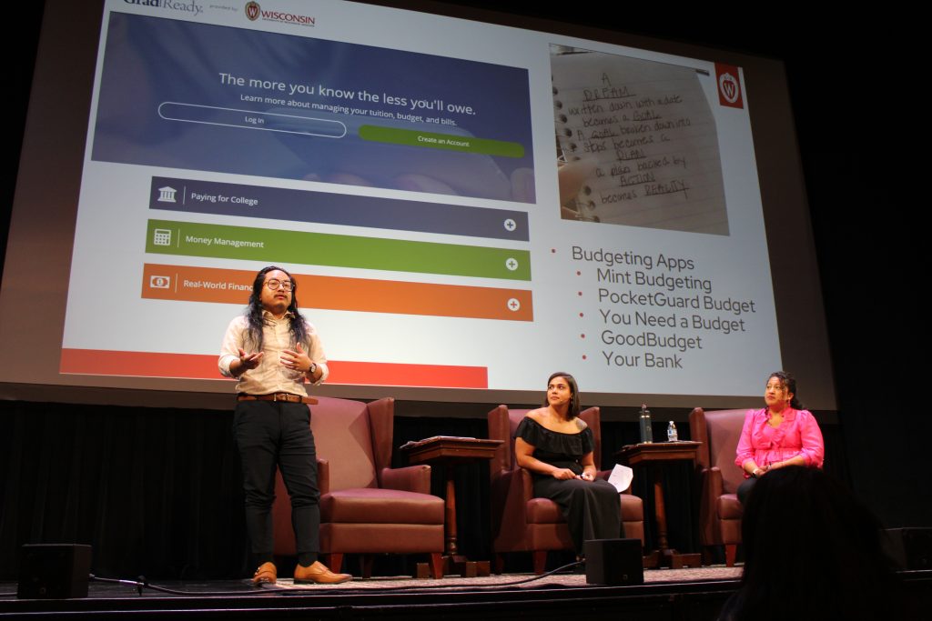Presentation with a standing speaker and two seated panelists on stage.