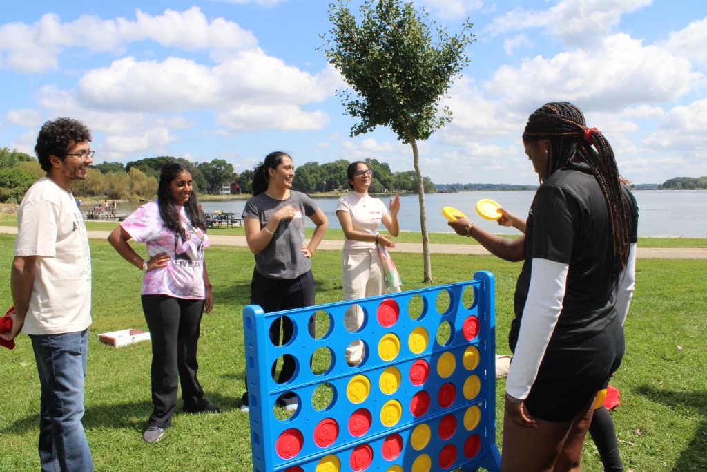 Group of people playing a large Connect Four game outdoors near water and trees.