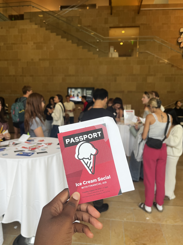 A hand shows a closeup of the cover of a Passport for the Financial Aid Ice Cream Social in a busy venue.