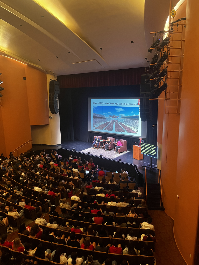 View of a filled auditorium listening to a panel with a screen presentation behind them.