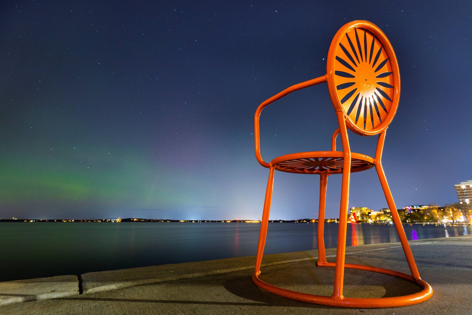 An orange Memorial Union Terrace chair in the foreground with the Northern Lights in the background.