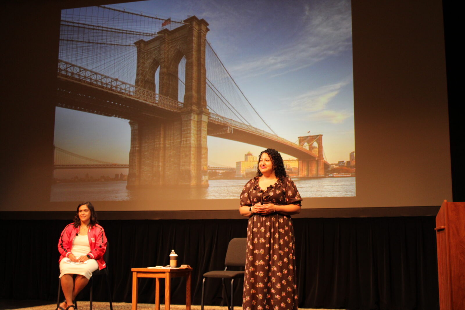 Joselyn presents in front of a screen showing the Brooklyn Bridge with MollyJo sitting closeby.